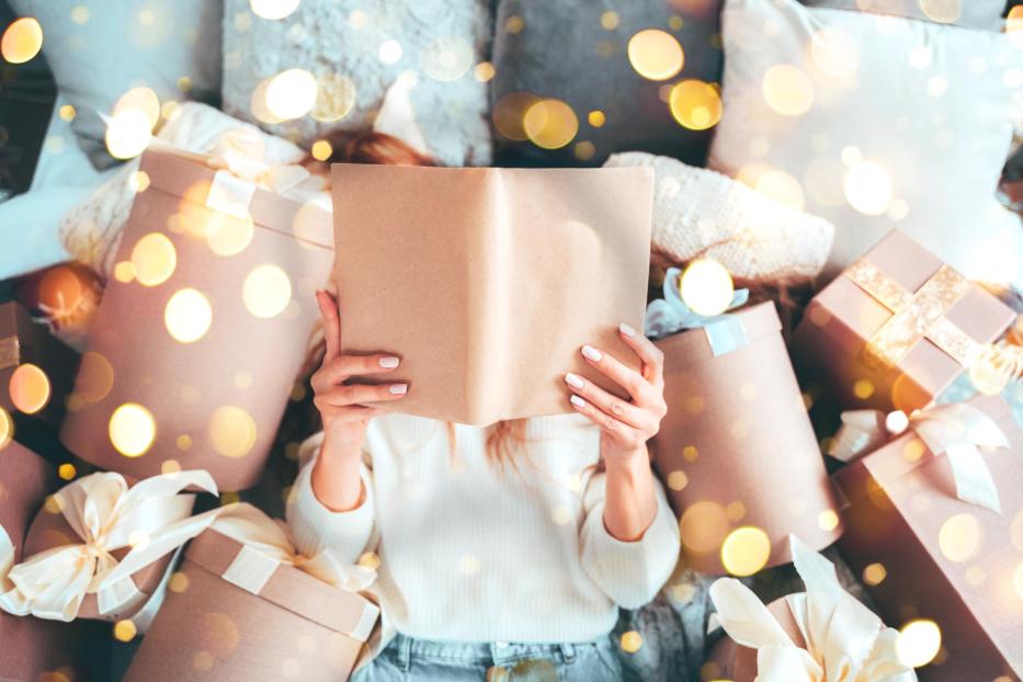 Beautiful woman is reading a book, laying between gift boxes, enjoying Christmas sale offers - Stock-Fotografie