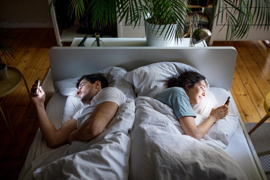 Man and woman lying against each other in bed and using their mobile phone. Couple ignoring each other and using their smart phones.