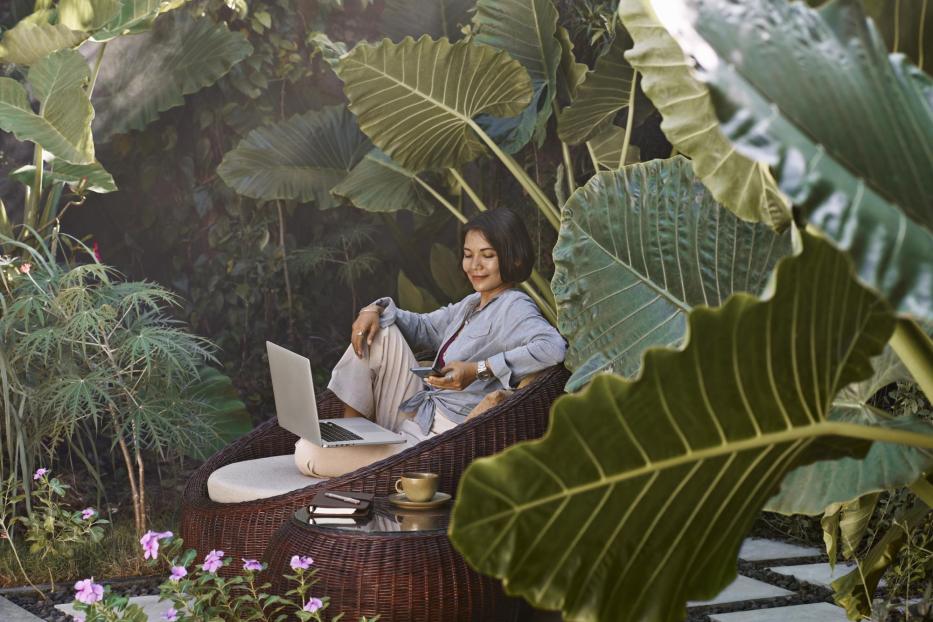 Asian woman working from home sitting in garden furniture surrounded by tropical plants, using laptop computer and mobile phone - Stock-Fotografie