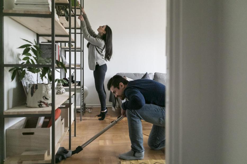 Man vacuuming floor while woman cleaning shelf at home - Stock-Fotografie