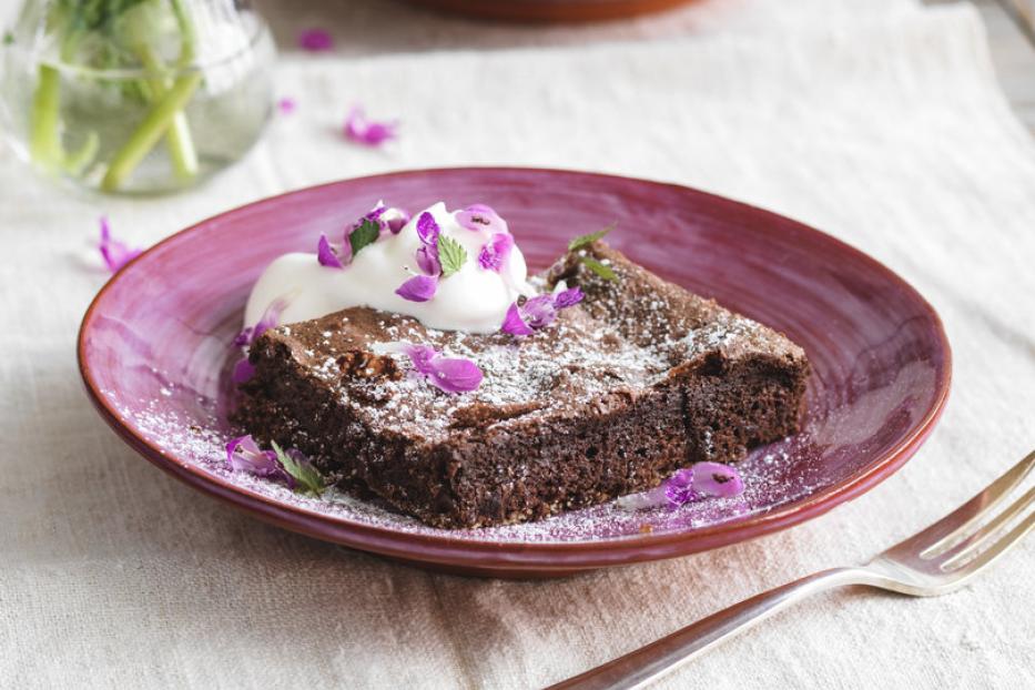 Ein Schokoladenbrownie mit Puderzucker, Sahne und lila Blüten auf einem Teller.