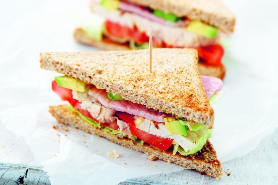 Zwei angeschnittene Sandwiches mit Hähnchen, Avocado, Tomaten und Salat auf einem Holzbrett.