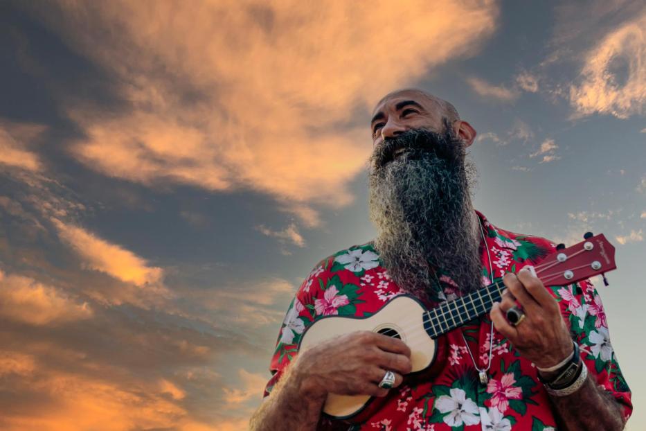 Rasierter Kopf reifer Mann mit langem Bart gekleidet mit Hawaiihemd spielt Ukulele mit einem dramatischen Himmel im Hintergrund - Stock-Fotografie