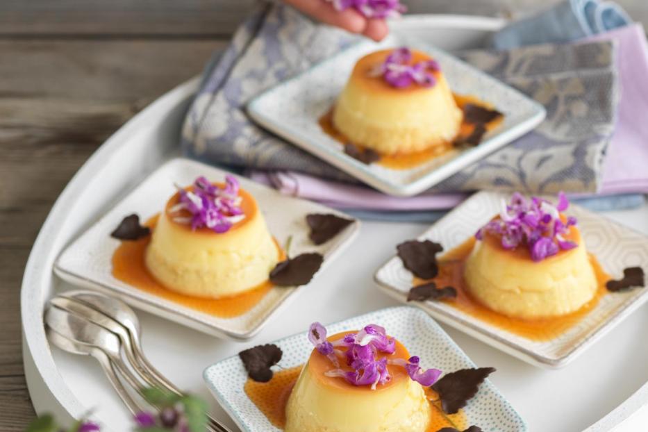 Vier Puddingförmchen mit Karamell und lila Blüten auf einem Tablett angerichtet.