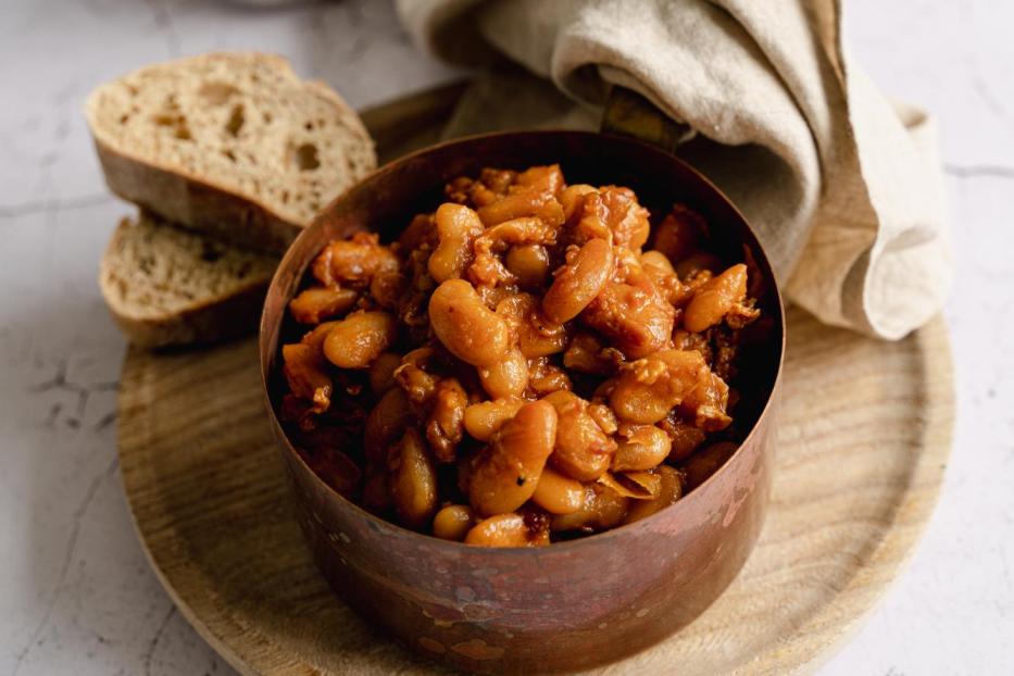 Ein Topf mit Baked Beans steht auf einem Holzteller, dazu Brot und ein Bier.