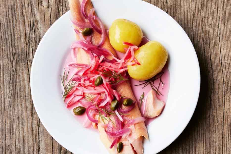 Ein Teller mit gebeiztem Lachs, Kartoffeln, roten Zwiebeln und Kapern auf einem Holztisch.