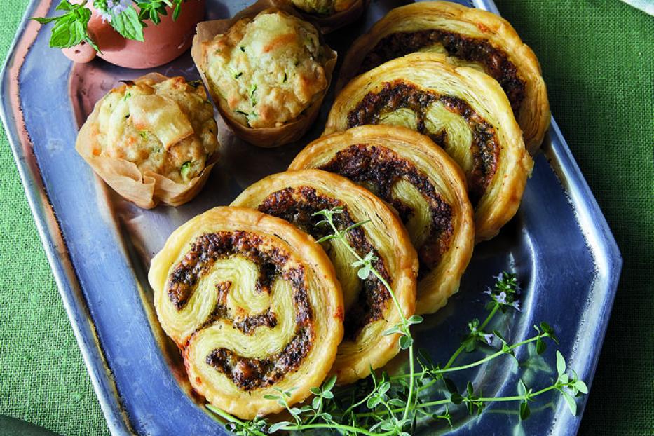 Ein silbernes Tablett mit Muffins, Blätterteigschnecken und einem kleinen Topf mit Kräutern.
