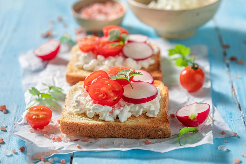 Zwei Toasts mit Hüttenkäse, Tomaten und Radieschen auf blauem Untergrund.