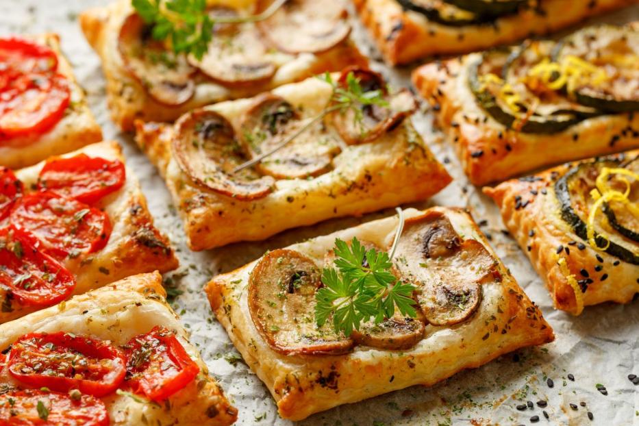 Blätterteig-Häppchen belegt mit Tomaten, Pilzen und Zucchini auf einem Backpapier.