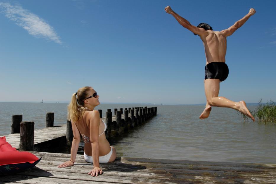 Ein Mann springt ins Wasser, während eine Frau auf einem Steg sitzt.