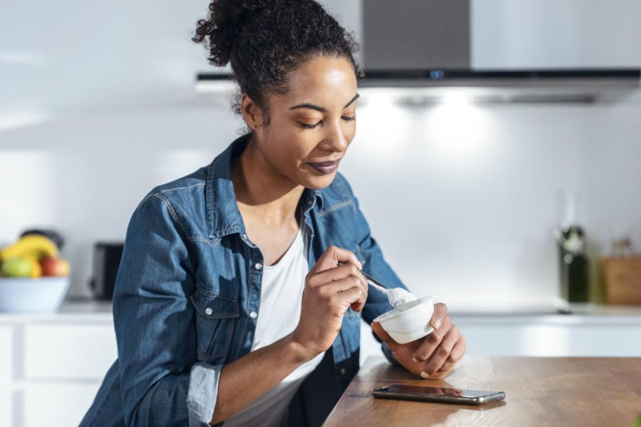 Woman eating yogurt sitting in kitchen at home - Stock-Fotografie