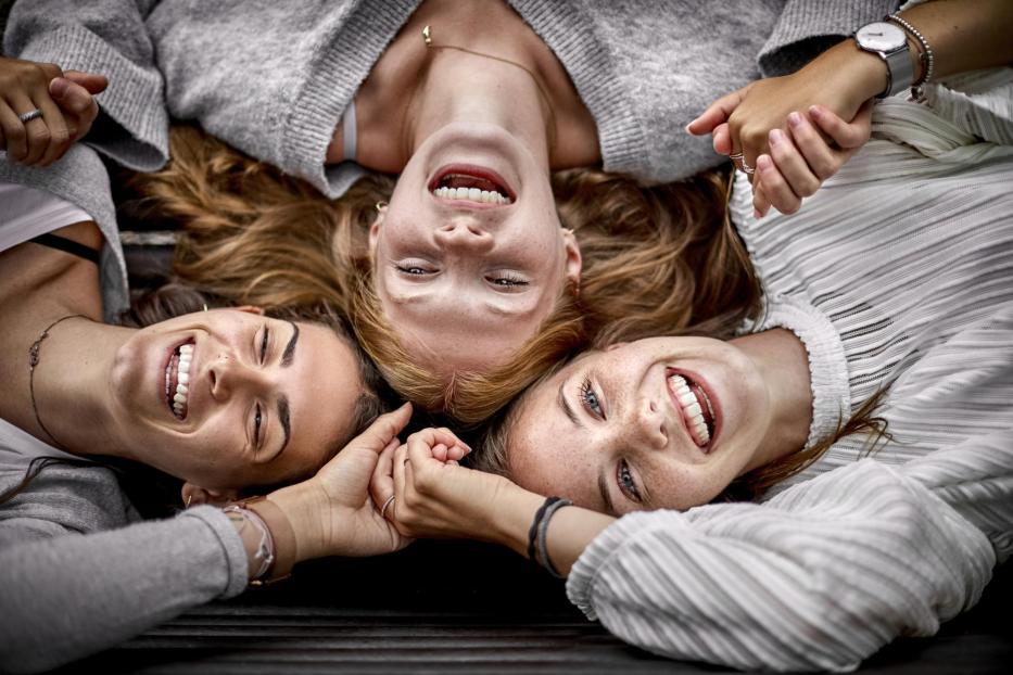 Three happy young women lying on a bench holding hands - Stock-Fotografie