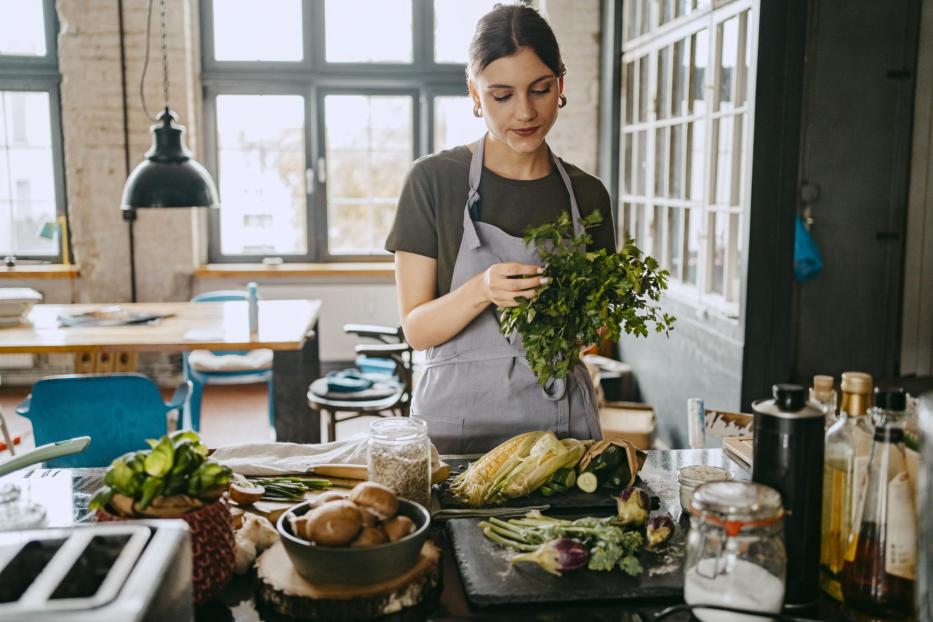 Female chef wearing apron doing quality check of cilantro standing in studio kitchen - Stock-Fotografie