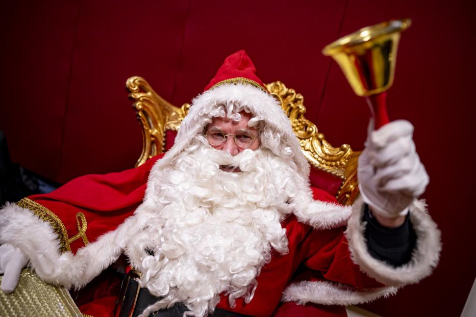 Weihnachtsmann mit Bart und Glocke in der Hand sitzt auf einem goldenen Thron.