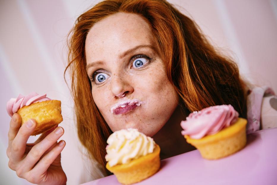 Eine rothaarige Frau mit Zuckerguss im Gesicht hält einen Cupcake und blickt mit großen Augen auf weitere Cupcakes.