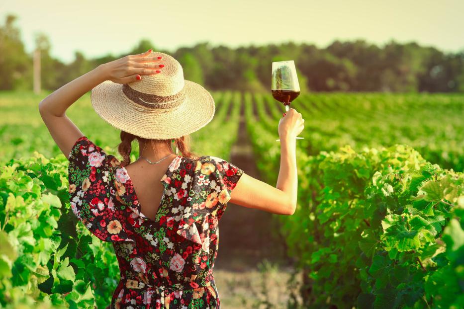 Frau im Sommerkleid und mit Sonnenhut hebt ein Weinglas mit Rotwein hoch, steht inmitten von Weinbergen