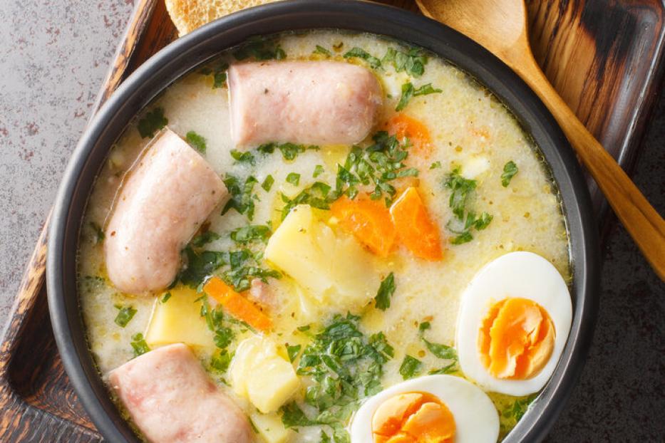 Polnische Suppe Zurek mit Weißwurst, Eiern, Kartoffeln und Karotten in einer schwarzen Schüssel, daneben ein Holzlöffel und Brot.