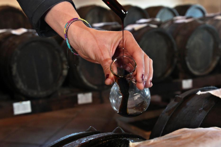 Hand mit Glasflasche und dunkler Flüssigkeit vor Fässern