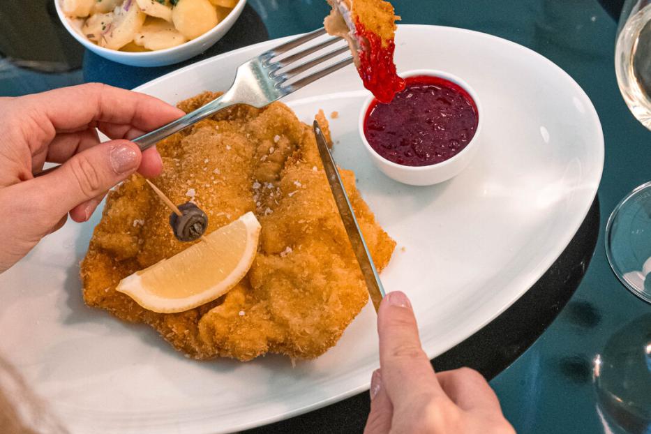 Eine Hand hält eine Gabel, worauf sich ein Schnitzelstück befindet. In der anderen ist ein Messer. Vor der Person liegt auf einem Teller ein schönes Wiener Schnitzel, garniert mit einer Zitronenspalte und einem Sardellenring.