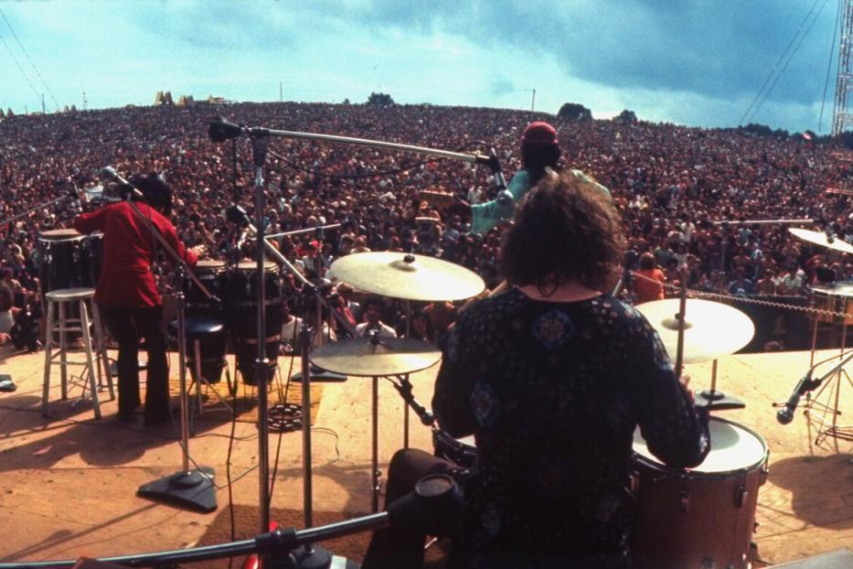 Eine Band auf der Bühne beim Woodstock Festival 1969. Von der Bühne blicken die Musiker inmitten eine Menschenmasse