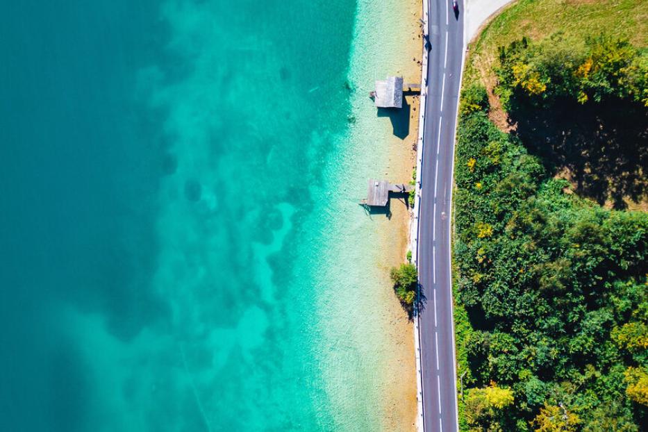 Eine Drohnenaufnahme vom Attersee: Eine Wiese mit Bäumen, dann eine Straße am Ufer und daran grenzt der klare Attersee in verschiedenen Blautönen.