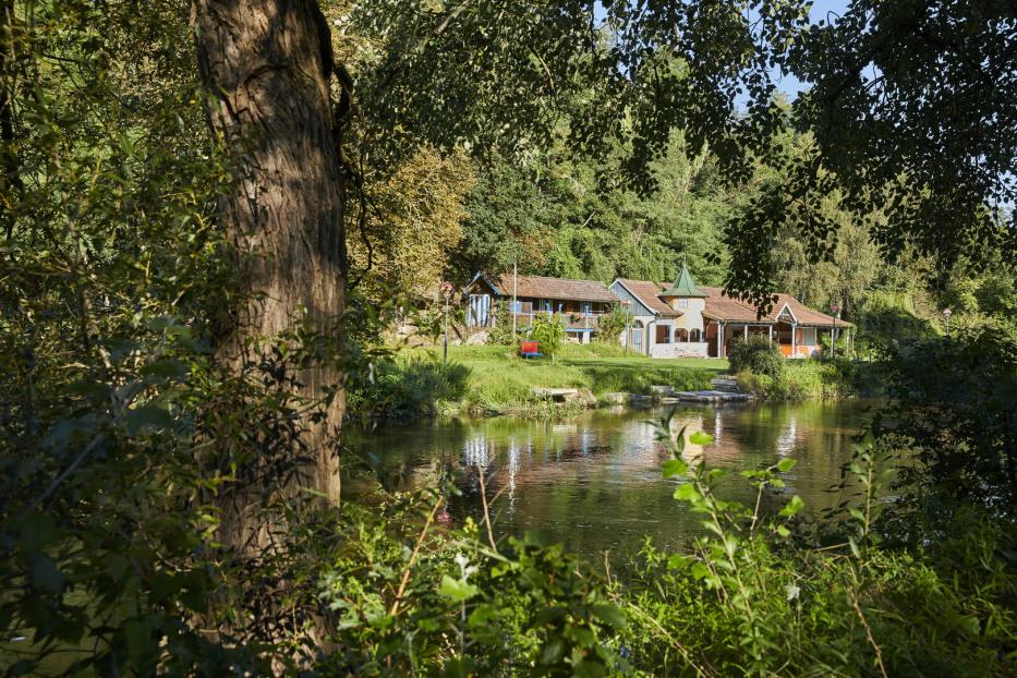 Grün-braunes Wasser, Bäume, eine Liegewiese und im Hintergrund ein Holzgebäude, das einst das Flussbad beherbergte