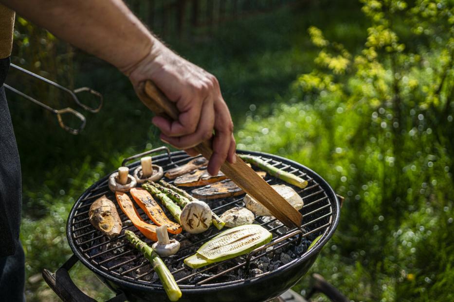 Kleiner Griller, auf dem Rost liegen Zucchini, Champignons, Spargel und Melanzani, ein Mann wendet das Gemüse mit einer Grillzange