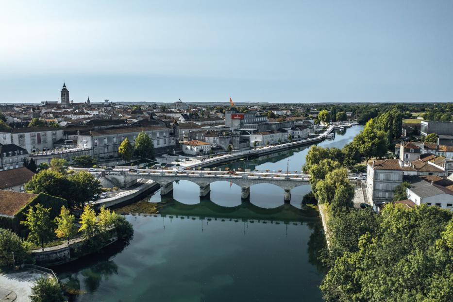 Eine Brücke über dem Fluss Charente. Am Ufer stehen Teile der Stadtmauer, ein großes Gebäude des Cognac-Herstellers Hennessey, im Hintergrund erhebt sich die Kirche.