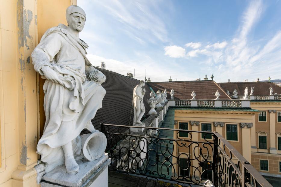 Steinfiguren schauen beim Balkon unterhalb der großen Uhr von oben auf den Ehrenhof des Schlosses Schönbrunn herab. Vor dem Balkon befindet sich ein Schmiedeisengitter, vor dem Dach eine steinerne Brüstung