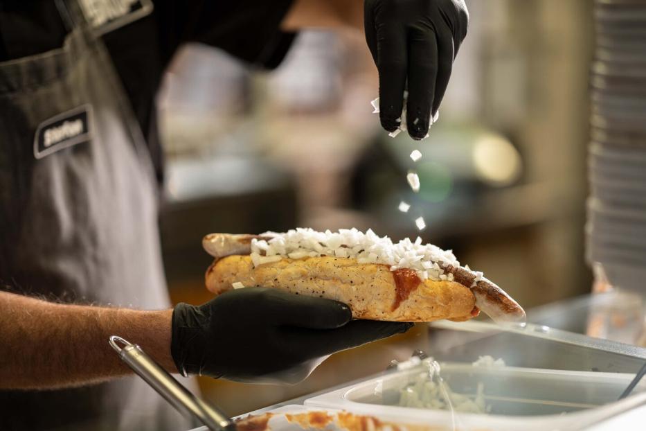 Eine Bosna entsteht: Hände in Handschuhen halten ein Baguette: Darin liegt eine Bartwurst, die Hand streut Zwiebeln darauf.