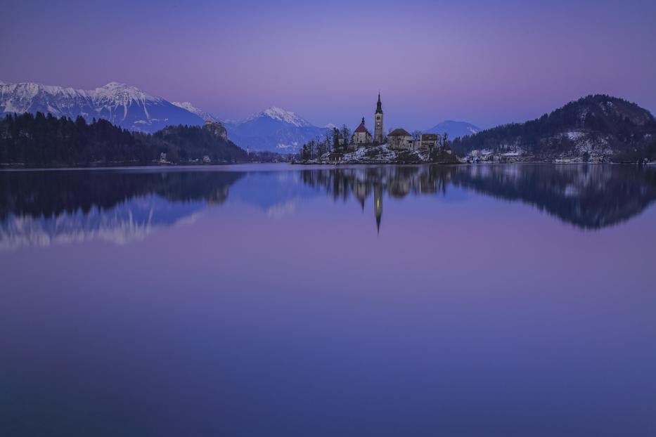 Eine Kircheninsel inmitten des Bleder Sees, aufgenommen am Abend. Rundherum stehen Hügeln und Berge