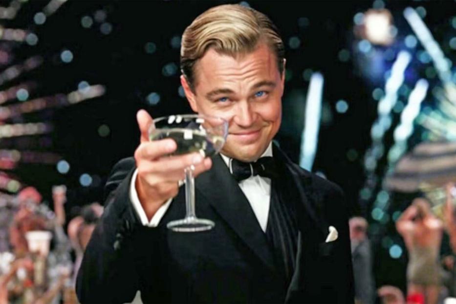 Der große Gatsby im Smoking, gespielt von Leonardo DiCaprio, prostet mit einer Champagnerschale. Im Hintergrund explodiert ein Feuerwerk.