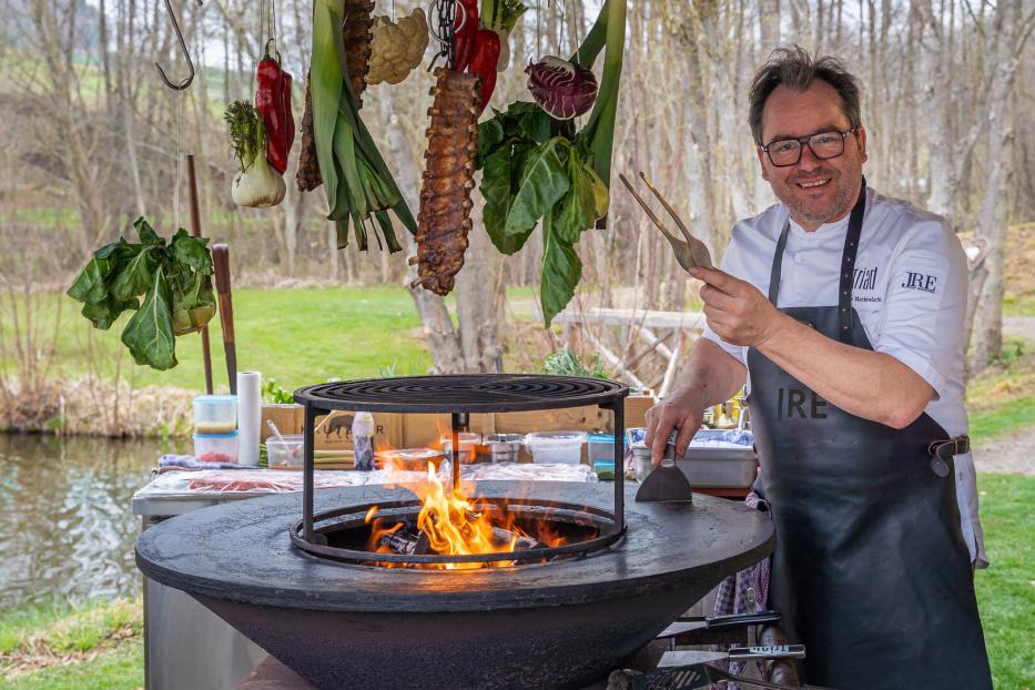 Uwe Machreich bei seinem Griller aus Platte und Feuerstelle. Darüber hängen Ripperl und Gemüse.