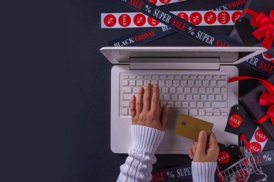 Person in weissem Pullover tippt auf einem Laptop und hält eine Kreditkarte in der anderen Hand; rundherum liegen Geschenkschachteln, ein kleiner Einkaufswagen und Bänder mit Aufschriften wie "Black Friday", "Sale" und "Super Sale" auf dunklem Hintergrund.