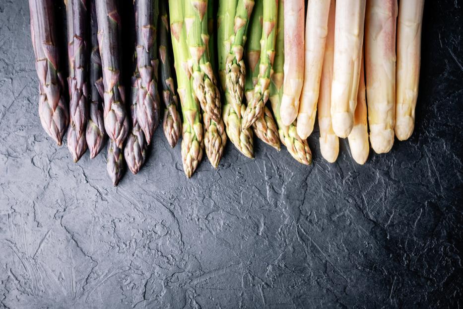 Drei Sorten Spargel, lila, grün und weiß, liegen nebeneinander auf dunklem Untergrund.