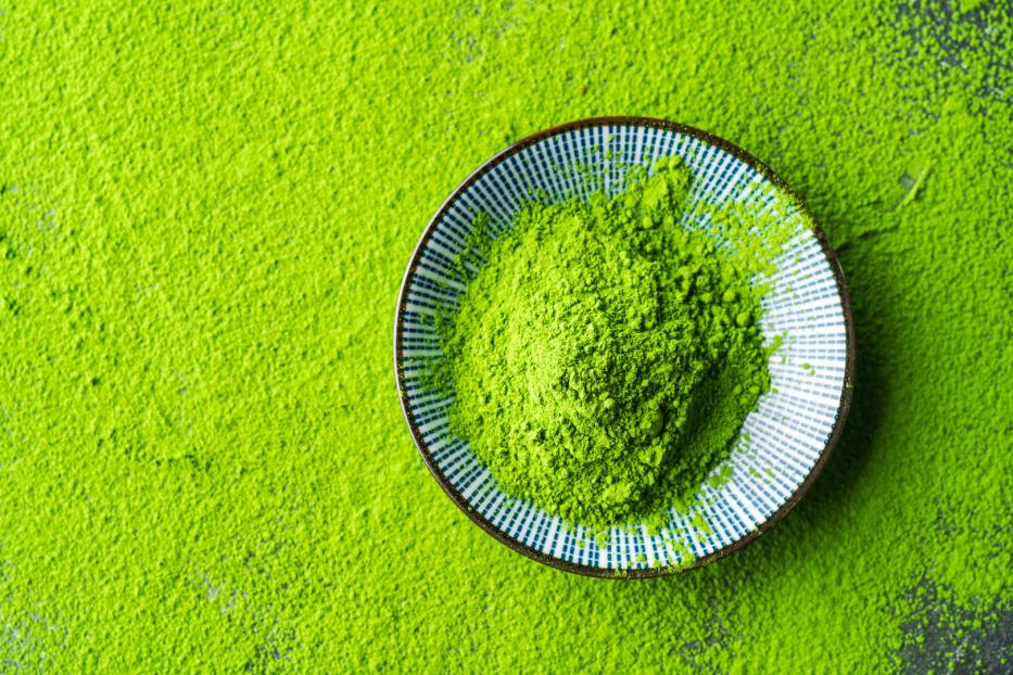 Eine kleine Schale mit leuchtend grünem Matcha-Pulver steht auf einer Fläche, die ebenfalls mit Pulver bedeckt ist.
