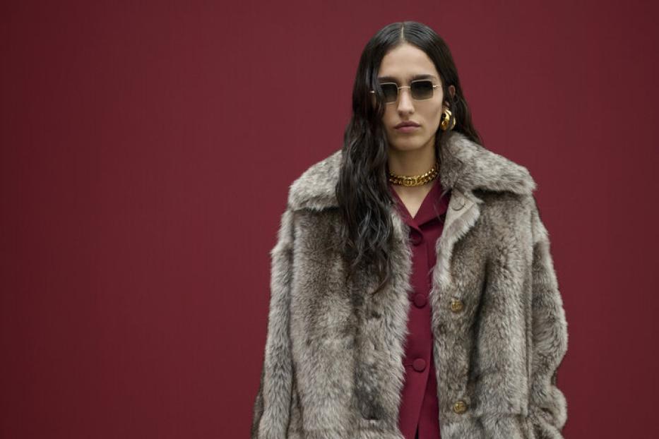Eine Frau mit Sonnenbrille, Pelzmantel und Handtasche vor einem roten Hintergrund.