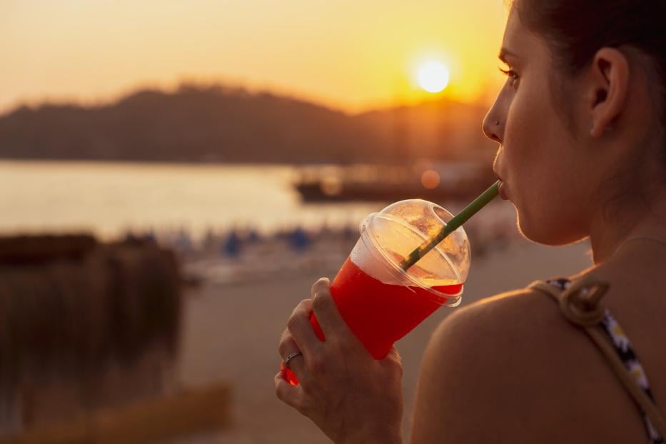 Eine junge Frau schlürft genüsslich einen Slushie im Sonnenuntergang an einem Seeufer