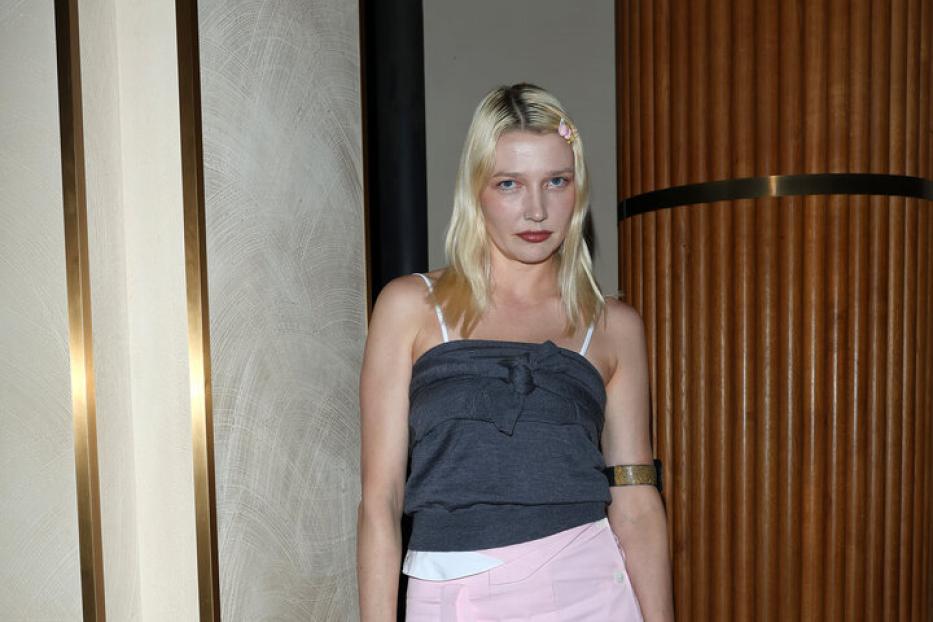 Eine Frau mit blonden Haaren trägt ein graues Top, einen rosa Rock und eine braune Handtasche von Miu Miu.