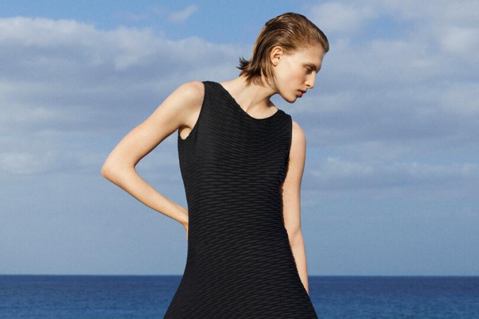 Eine Frau in einem schwarzen Kleid und Sandalen steht vor dem Meer.