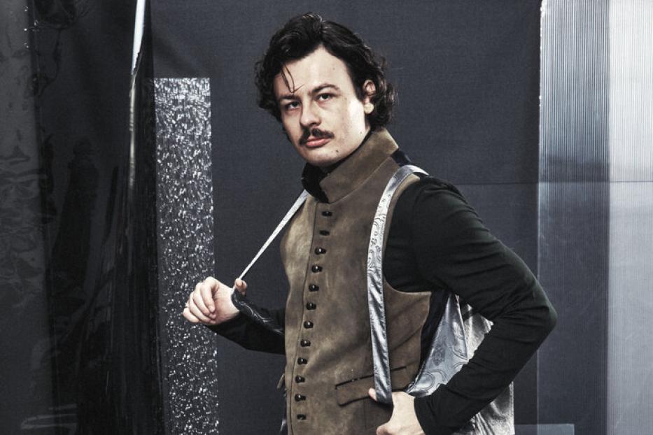 Ein Mann mit Schnurrbart trägt eine Lederhose, eine Weste und Stiefel vor einem grauen Hintergrund.