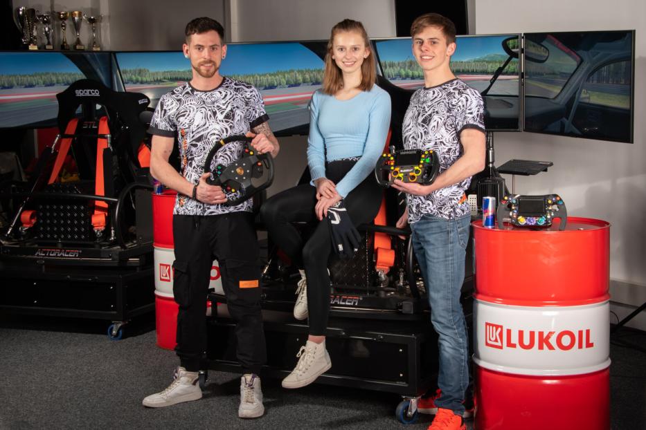 Zwei junge Männer und ein junges Mädchen posieren unter anderem mit einem Autolenkrad vor einem Rennsimulator.