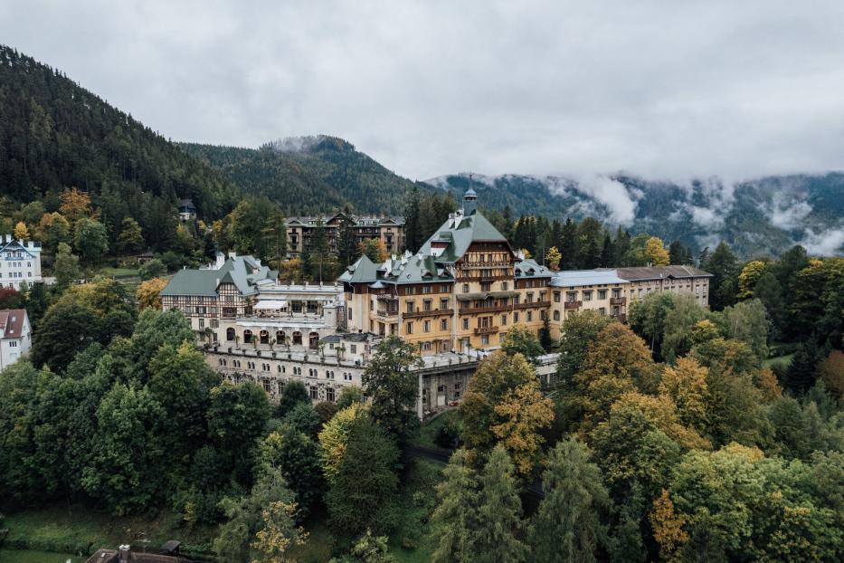 Das große Südbahnhotel mit seinen verschiedenen Trakten und großem Balkon. Es thront auf einem bewaldeten Hang. Im Hintergrund erheben sich Berge, Nebel lichtet sich