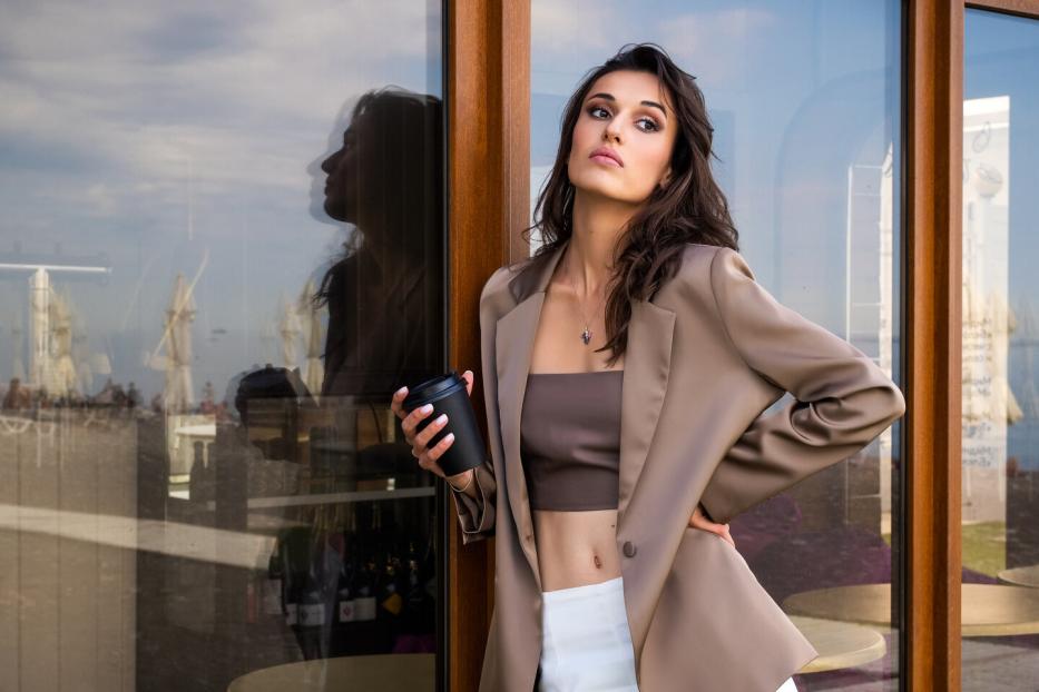 Ein attraktives Model posiert vor einem Café mit einer Tasse Kaffee in der Hand und trägt ein Bandeau-Top, eine weiße Hose und ein Sakko.