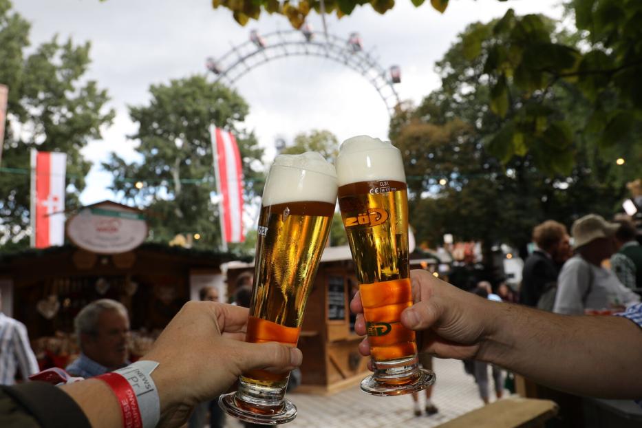 Zwei Hände mit Biergläsern, die gerade zusammenstoßen, im Hintergrund sieht man das Wiener Riesenrad