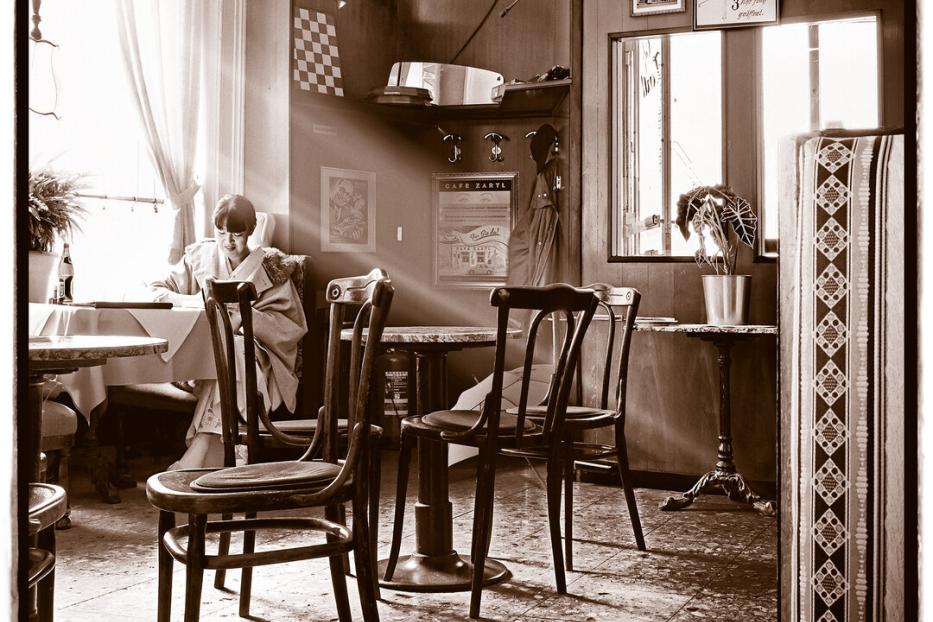 Eine Frau sitzt im Kaffeehaus am Fenster, Sonnenlicht fällt rein. Sie liest Zeitung.