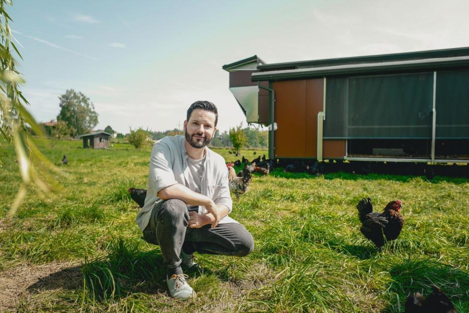 Ein Mann hockt auf einer Wiese mit Hühnern vor einem mobilen Hühnerstall.