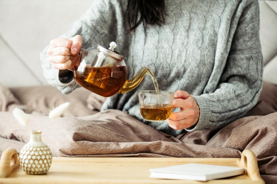 Eine Person gießt Tee aus einer Glaskanne in eine Tasse auf einem Tablett im Bett.