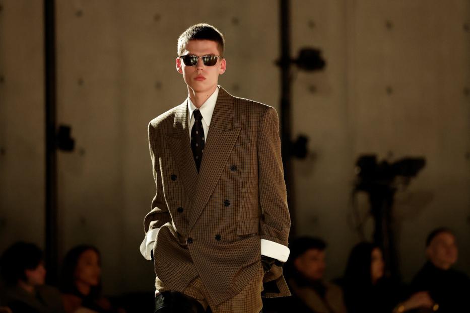 Juger Mann mit Zweireiher und Sonnenbrille am Laufsteg. Die Hände hat er in der Hosentasche.