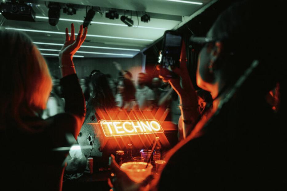 Blick vom DJ-Pult auf den Innenraum. Menschen haben die Hände in der Höhe und tanzen, eine Person fotografiert.. Auf einem Leuchtschild steht Techno.